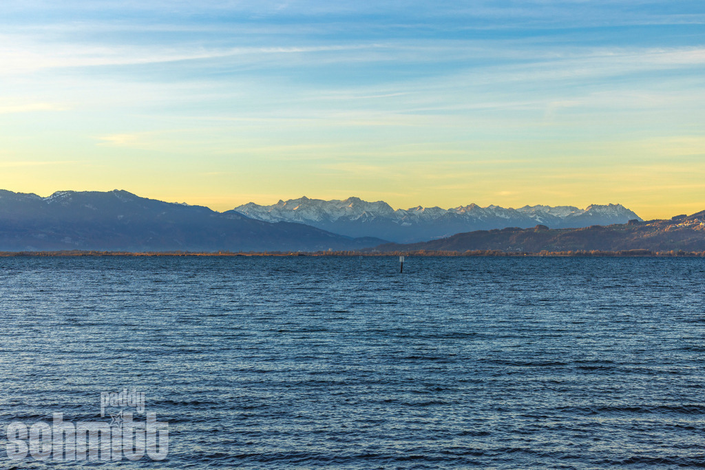 Alpenpanorama vor dem Bodensee am frühen Morgen | Am frühen Morgen liegt der Bodensee ruhig vor der beeindruckenden Kulisse der Alpen. Das sanfte Licht des Tages lässt die Bergsilhouette besonders klar hervortreten und verleiht dem weiten Panorama eine ruhige, zeitlose Atmosphäre. - Realisiert mit Pictrs.com