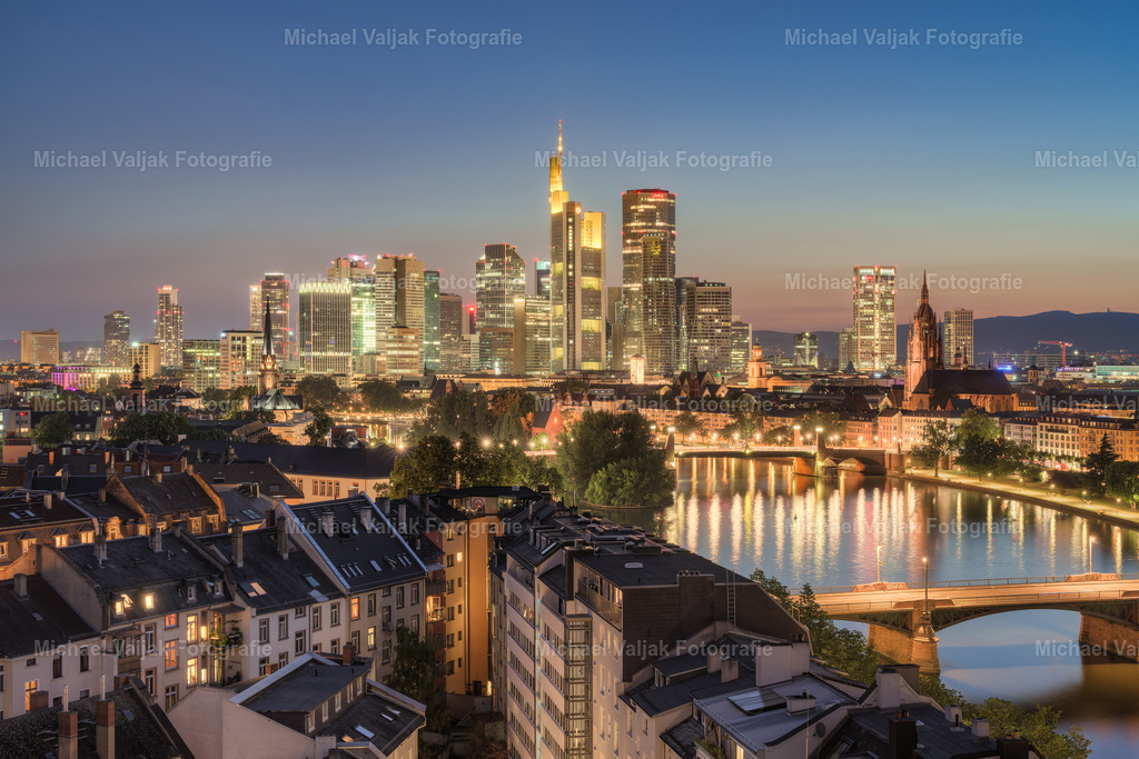 Blick über Frankfurt am Abend | Die Stadt zeigt sich im Abendlicht klar und strukturiert. Die Hochhäuser stehen ruhig vor dem dunkler werdenden Himmel, ihre Fenster beginnen zu leuchten. Von oben erkennt man die regelmäßige Anordnung der Straßen und Gebäude – eine Mischung aus Wohn- und Bürovierteln, dicht und funktional. - Realisiert mit Pictrs.com