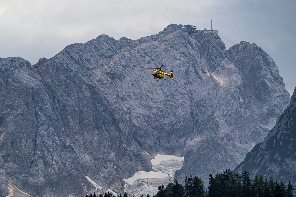 Einsatz vor gigantischer Kulisse: Christoph 74 an der Zugspitze | Dramatische Dynamik und alpine Erhabenheit in einem Bild: Der intensiv gelbe Rettungshubschrauber Christoph 74 (Kennung D-HYAE, Typ Airbus Helicopters H145) der ADAC Luftrettung aus Murnau schwebt vor den gewaltigen, schroffen Felswänden des Wettersteingebirges bei Garmisch-Partenkirchen. Im Hintergrund ragt das Zugspitzmassiv mit der Gipfelstation der Zugspitzbahn und den verbliebenen Schneefeldern des Höllentalferners eindrucksvoll empor. Eine fesselnde Aufnahme voller Hoffnung, Präzision und bergsteigerischer Realität. - Realisiert mit Pictrs.com