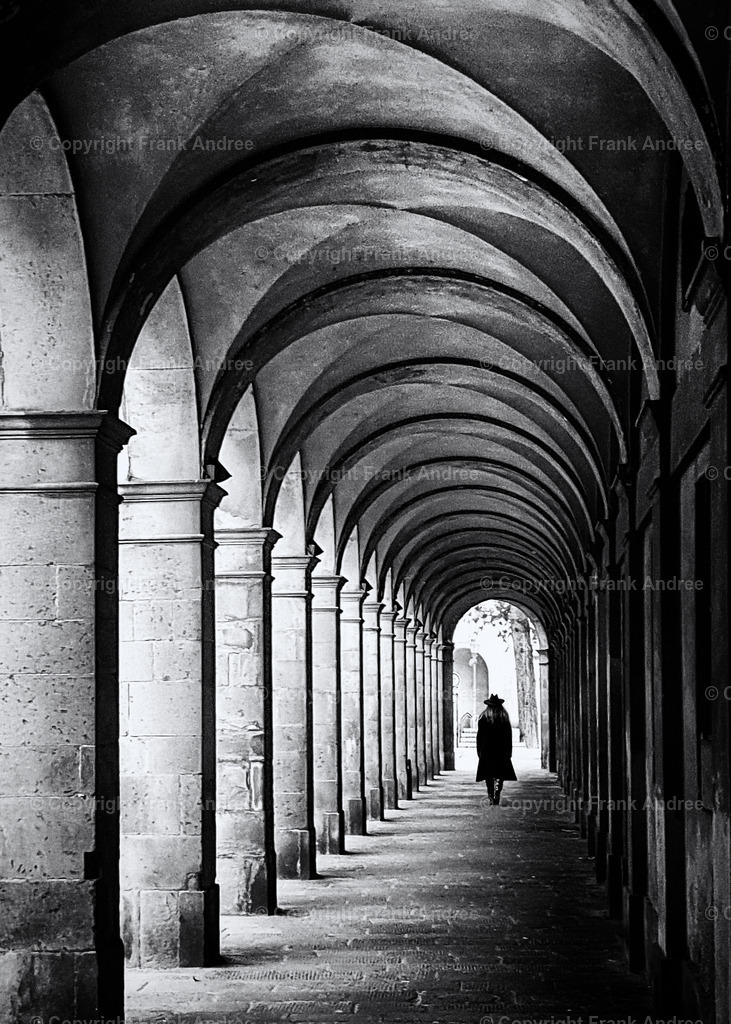 Lucca | Eine Frau mit Mantel und Hut geht allein durch eine Arkade in der Altstadt von Lucca. Schwarz weiß Fotografie aus der Toskana Italien. - Realisiert mit Pictrs.com
