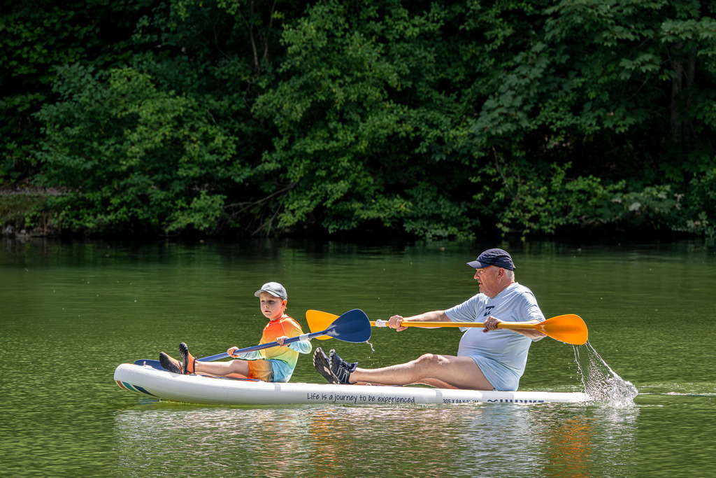 Gemeinsam auf Kurs: Sommerlicher Paddelspaß auf dem Riessersee | Ein Mann und ein kleiner Junge gleiten mit spritzenden Paddelschlägen auf einem weißen Stand-up-Paddle-Board über das ruhige, grün schimmernde Wasser des Riessersees. Während der Junge vorn konzentriert mitpaddelt, sorgt der Mann hinten für den nötigen Antrieb und die richtige Richtung. Vor der dichten Kulisse des tiefgrünen Uferwaldes vermittelt das Motiv echtes Teamwork, gemeinsame Abenteuerfreude und die unbeschwerte Leichtigkeit eines perfekten Sommertages im Garmisch-Partenkirchener Land. - Realisiert mit Pictrs.com