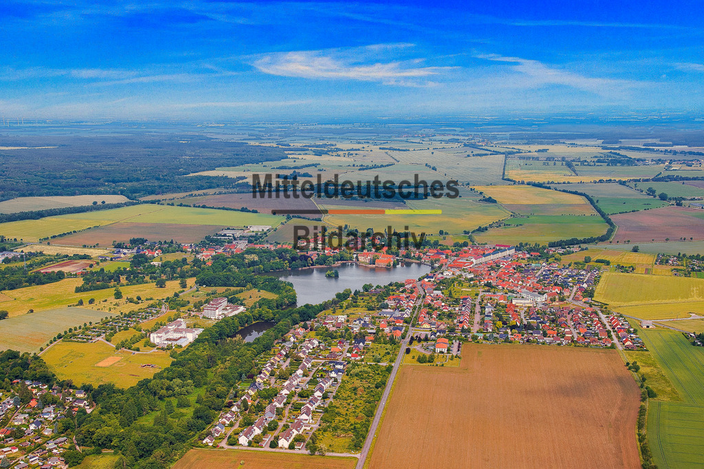 Flechtingen-See-Schloß-Börde-Sachsen-Anhalt_3689 | Die Luftaufnahme zeigt Flechtingen mit dem See als prägendem Bestandteil des Ortsbildes. Wohnbebauung, Grünflächen und Wasser bilden aus der Vogelperspektive eine charakteristische Einheit. In der weiteren Umgebung sind außerdem Gewerbeflächen und der Steinbruch zu erkennen, die eine andere Seite der Region dokumentieren. - Realisiert mit Pictrs.com