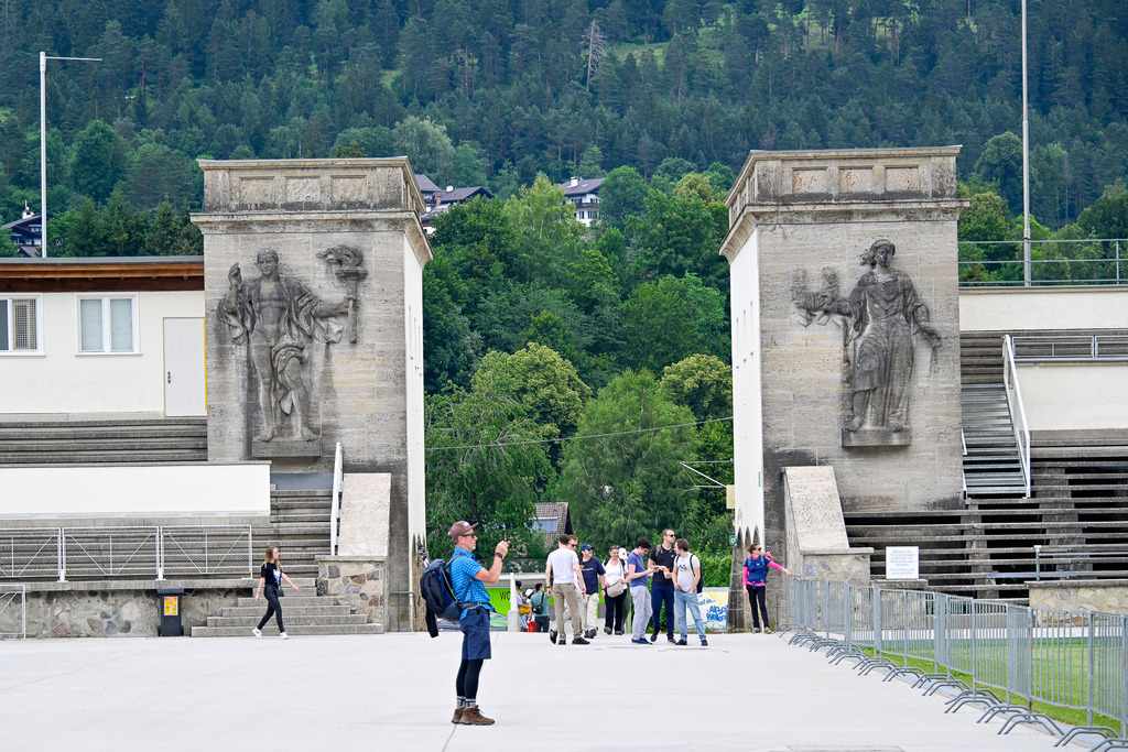 Steinerne Zeugen der Geschichte: Das Erbe von Garmisch-Partenkirchen | Ein Ort, an dem die Zeit spürbar stillsteht: Die monumentalen Steintürme des historischen Olympia-Skistadions in Garmisch-Partenkirchen ragen eindrucksvoll empor. Die klassischen Reliefs, die eine Fackelträgerin und einen Athleten zeigen, strahlen eine ehrwürdige Melancholie aus. Vor dieser Kulisse fängt die Aufnahme das lebendige Treiben von heute ein – Besucher, die den geschichtsträchtigen Platz erkunden und fotografieren. Im Hintergrund erheben sich die dicht bewaldeten Hänge der Alpen als zeitlose, grüne Wächter. Ein Bild, das die Brücke zwischen der Historie des Sports und der Gegenwart schlägt. - Realisiert mit Pictrs.com