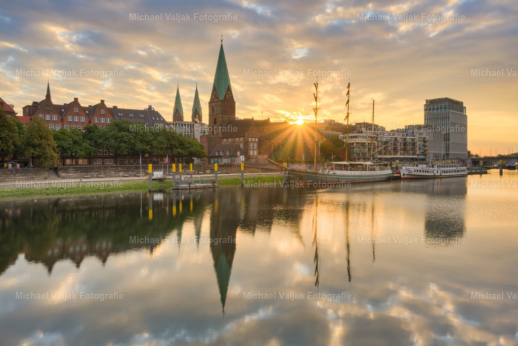 Sonnenaufgang an der Schlachte in Bremen | Kurz nach Sonnenaufgang erscheint die Sonne über den Dächern der Altstadt und bildet einen klaren Sonnenstern am Himmel. Die Gebäude entlang der Weser spiegeln sich im Wasser und erzeugen eine ausgewogene Komposition aus Licht und Struktur. Am Steg liegt das Segelschiff Alexander von Humboldt, eingebettet in die morgendliche Szenerie. Die Perspektive verbindet städtische Architektur mit maritimen Elementen in einem Moment stiller Klarheit. - Realisiert mit Pictrs.com