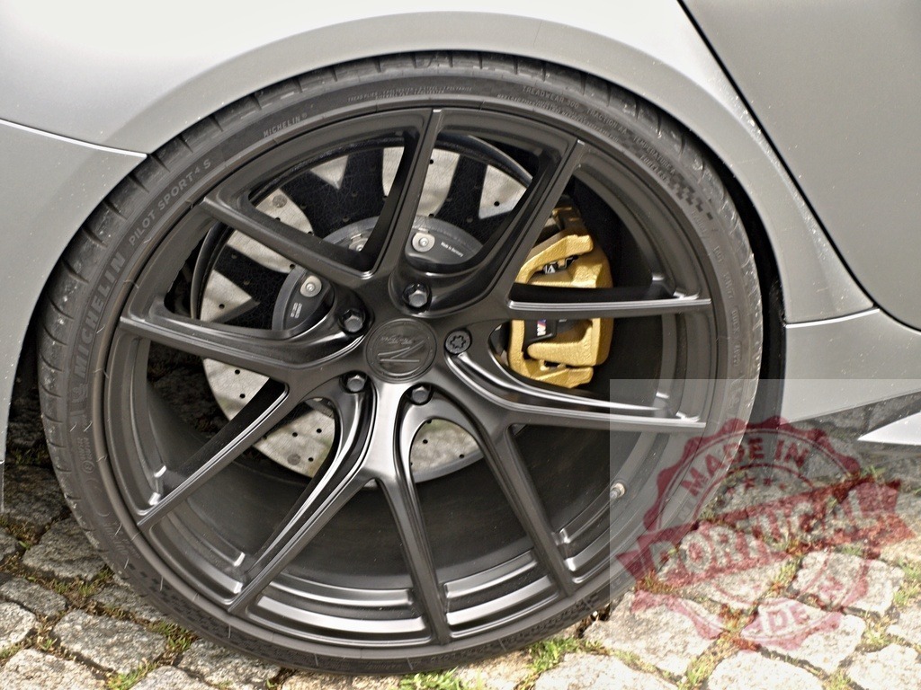 Performance car wheel with Michelin tire and gold brake caliper | Car wheel with black alloy rim, gold brake caliper, and perforated disc brake on cobblestone - Realized with Pictrs.com