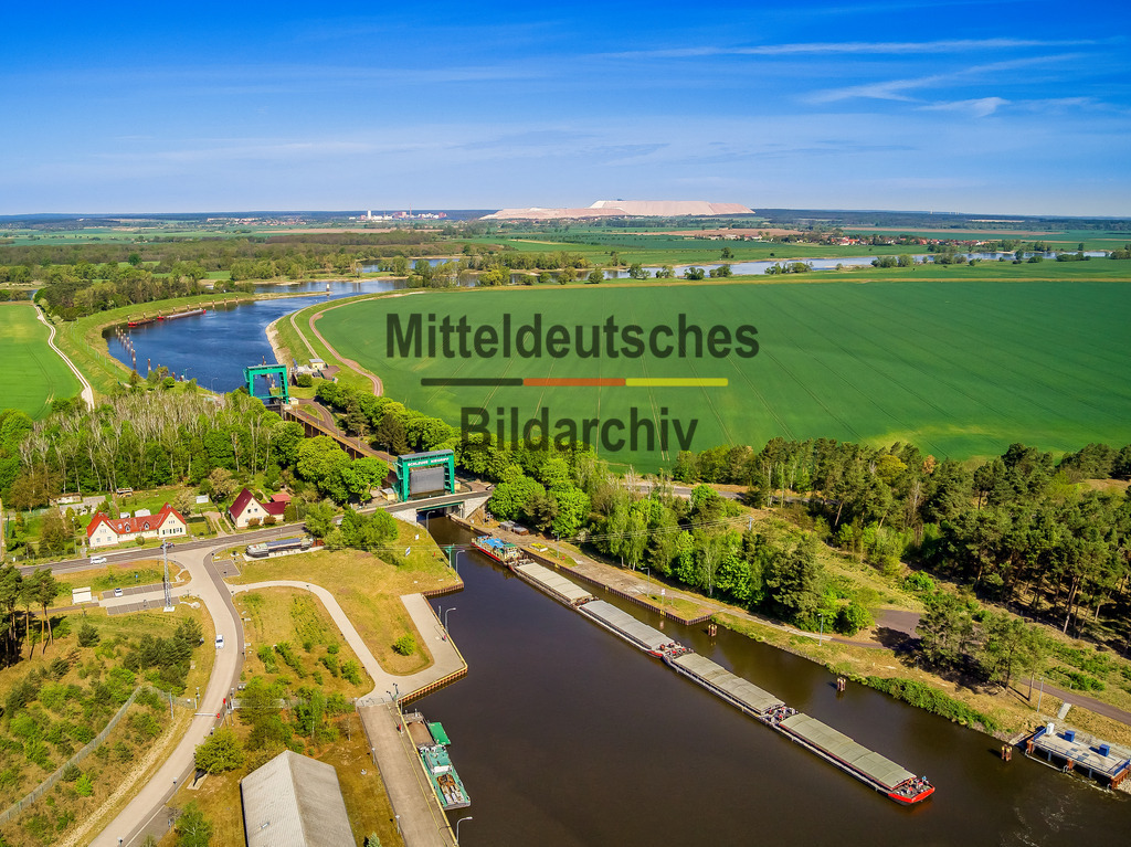 Luftbild_Schleuse_Niegripp_Schiff_Kaliberg_LR-0058 | Das Luftbild zeigt die Schleuse Niegripp aus einer Drohnenperspektive. Eingebettet in die weite Landschaft des Elbe-Havel-Kanals verlässt ein Schubverband die Schleusenkammer und verdeutlicht die Bedeutung der Anlage für die Binnenschifffahrt. Aus der Vogelperspektive werden die klaren Strukturen des Schleusenbauwerks sowie das Zusammenspiel von Wasserstraße, Technik und Landschaft besonders eindrucksvoll sichtbar.Die Schleuse Niegripp wurde 1938 in Betrieb genommen und zählt zu den wichtigsten Bauwerken des Elbe-Havel-Kanals. Die Schleusenkammer ist 225 Meter lang und 12 Meter breit und ermöglicht die Passage moderner Güterschiffe und Schubverbände. Mit einem Hub von rund 5,5 Metern gleicht die Schleuse den Höhenunterschied zwischen den Kanalabschnitten aus und gewährleistet einen leistungsfähigen Schiffsverkehr auf einer der bedeutendsten Wasserstraßen Ostdeutschlands.Die Drohnenaufnahme vermittelt einen eindrucksvollen Blick auf die Dimensionen der Schleusenanlage und die präzise Abwicklung eines Schleusenvorgangs. Der ausfahrende Schubverband unterstreicht die wirtschaftliche Bedeutung der Binnenwasserstraße und macht die Schleuse Niegripp zu einem markanten Beispiel moderner Wasserbau- und Verkehrstechnik. - Realisiert mit Pictrs.com