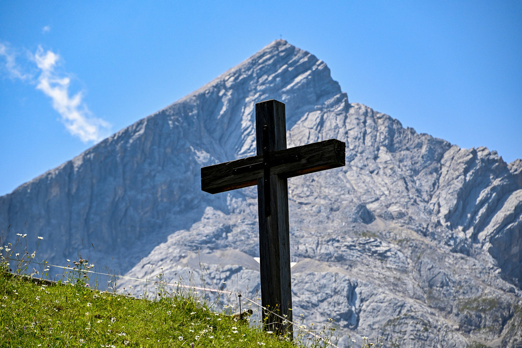 Alpen-Majestät und Gipfel-Glaube | Wie ein stiller Wächter steht das verwitterte Holzkreuz auf dem grünen Hang und blickt empor zur majestätischen Alpspitze. Der mächtige Gesteinskoloss, ein Wahrzeichen der bayerischen Alpen, ragt mit seiner charakteristischen Pyramidenform unbezwingbar in den strahlend blauen Himmel empor. Ein Hauch von Unendlichkeit liegt in der Luft, unterstrichen von den zarten Schleierwolken. Dieses Foto ist eine Hommage an die rohe Schönheit der Natur und den tiefen Glauben, der Wanderer und Bergsteiger auf ihrem Weg zum Gipfel begleitet. - Realisiert mit Pictrs.com