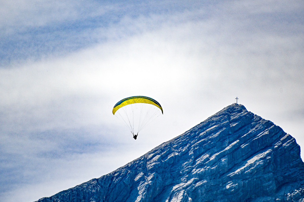 Gipfelglück und grenzenlose Freiheit | Ein Moment, der den Atem stocken lässt: Ein Paraglider schwebt mit seinem leuchtend gelben Schirm majestätisch über der Alpspitze. Unter ihm die gewaltige, schroffe Felswand, gekrönt vom markanten Gipfelkreuz. Diese Aufnahme fängt die unendliche Sehnsucht nach Freiheit und die faszinierende Demut vor der Erhabenheit der Bergwelt ein. Der Kontrast zwischen dem filigranen Flieger und dem massiven Bergmassiv sowie der sanfte Übergang vom klaren Blau zu den lockeren Wolken lassen den Betrachter die kühle Bergluft und das Adrenalin des Fliegers förmlich spüren. Ein Symbol für den Mut, die Grenzen zu überwinden und den Traum vom Fliegen zu leben. - Realisiert mit Pictrs.com