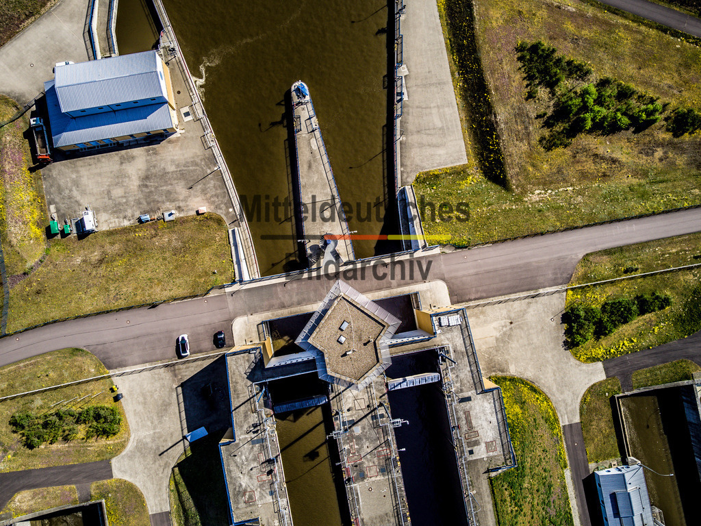 Schleuse Hohenwarthe-0123 | Das Luftbild zeigt die Schleuse Hohenwarthe aus einer senkrechten Drohnenperspektive. Der Blick direkt von oben macht die klare geometrische Anordnung der Schleusenkammer, der Sparbecken und der technischen Anlagen besonders eindrucksvoll sichtbar. Die symmetrische Bauweise und die präzise Linienführung verleihen der Anlage eine außergewöhnliche grafische Wirkung, die sich erst aus der Vogelperspektive vollständig erschließt.Die Schleuse Hohenwarthe wurde 2003 in Betrieb genommen und ist Bestandteil des Wasserstraßenkreuzes Magdeburg. Die beiden Schleusenkammern sind jeweils 190 Meter lang und 12,50 Meter breit. Mit einer Fallhöhe von bis zu 19 Metern zählt die Anlage zu den größten Sparschleusen Europas. Durch die insgesamt acht Sparbecken werden bei jedem Schleusenvorgang bis zu 60 Prozent des benötigten Wassers eingespart – ein wesentlicher Beitrag zu einem wirtschaftlichen und nachhaltigen Betrieb der Wasserstraße.Die senkrechte Drohnenaufnahme verdeutlicht die beeindruckende Präzision moderner Wasserbautechnik. Die geometrischen Formen der Schleusenanlage, kombiniert mit ihrer ingenieurtechnischen Funktion, machen die Schleuse Hohenwarthe zu einem der bedeutendsten Bauwerke des deutschen Binnenwasserstraßennetzes. - Realisiert mit Pictrs.com