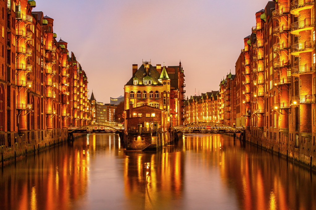 Goldene Stunde in der Speicherstadt - Hamburg leuchtet | Ein echter Klassiker der Architektur- und Städtefotografie: Dieses stimmungsvolle Wandbild zeigt die Hamburger Speicherstadt in ihrer schönsten Form – in der goldenen Stunde bei Abenddämmerung. Die warmen Lichtreflexionen auf dem Wasser, die historischen Backsteinfassaden und das markante Wasserschloss in der Mitte erzeugen eine einmalige Atmosphäre zwischen Tradition und Moderne. Ein beeindruckendes Kunstwerk für alle, die Hamburg lieben – oder urbane Eleganz und Lichtstimmung in ihren Räumen genießen möchten.