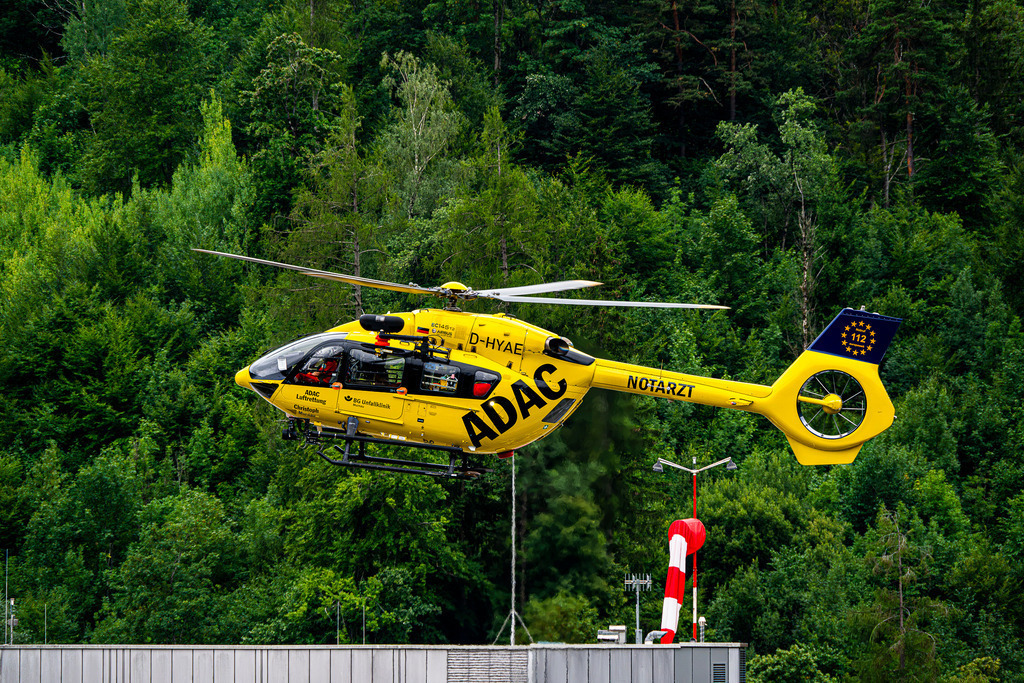 Gelber Engel vor grüner Kulisse: Christoph Murnau im Einsatz | Ein leuchtend gelber ADAC-Rettungshubschrauber der Type H145 (Kennung D-HYAE, „Christoph Murnau“) schwebt im tiefen Präzisionsflug vor dem dicht bewaldeten Steilhang. Im Vordergrund markiert ein rot-weißer Windsack den Hubschrauberlandeplatz. Die Aufnahme fängt das dynamische Zusammenspiel aus modernster Flugtechnik und der unberührten Natur der Alpenregion ein. Sie vermittelt ein starkes Gefühl von Schutz, Einsatzbereitschaft und Hoffnung – ein eingespieltes Rettungssystem, das in Notfällen schnellste Hilfe aus der Luft garantiert. - Realisiert mit Pictrs.com