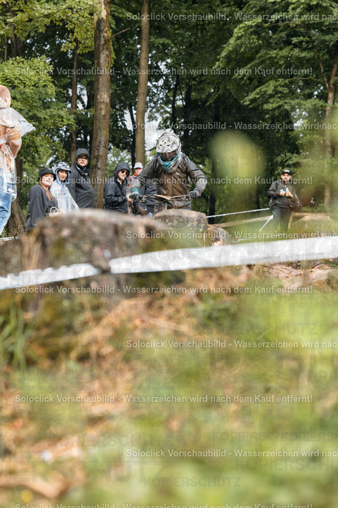Mountainbike-Action im Wald | Ein Mountainbiker in voller Ausrüstung springt über eine Rampe in einem bewaldeten Gelände. Im Hintergrund sind Zuschauer zu sehen, die das Ereignis beobachten. Die Szene vermittelt ein Gefühl von Action und Abenteuer. - Realisiert mit Pictrs.com