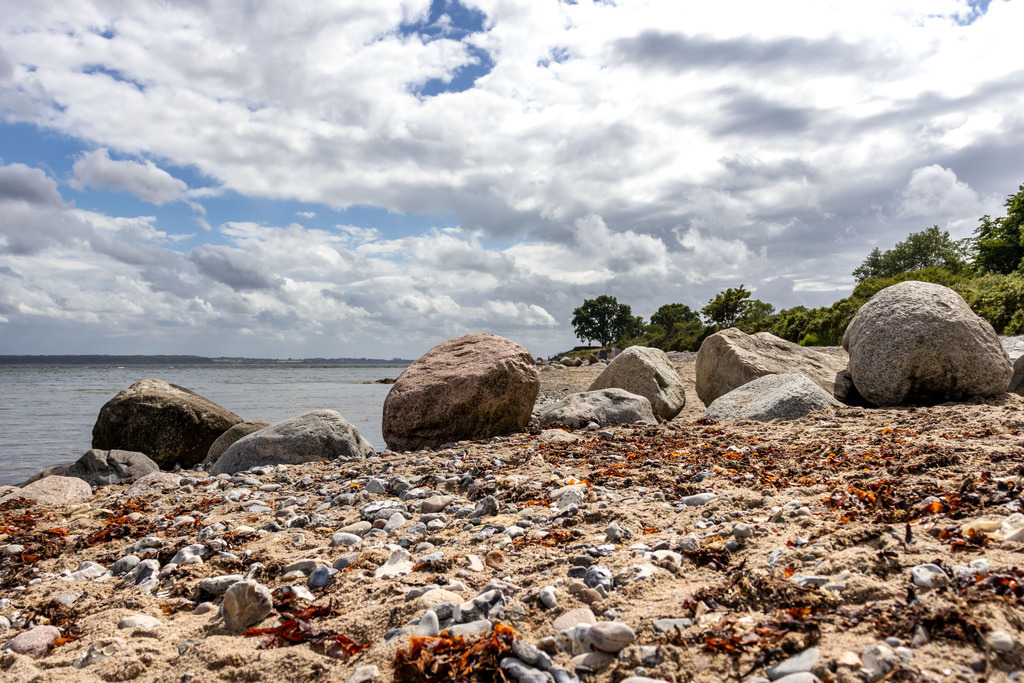 Wandbild: Strand in Langholz im Sommer | Dieses Wandbild im Querformat zeigt den Strand in Langholz im Sommer. In der Mitte des Bilds befindet sich eine Steinmole. Am blauen Himmel sind zahlreiche Wolken zu sehen.  - Realisiert mit Pictrs.com