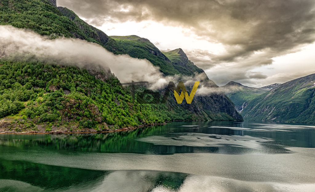 Dunkle Wolken im Geiranger Fjord------Unwetter in Norwegen | Das Bild zeigt den Geirangerfjord in Norwegen. Er ist einer der bekanntesten Fjorde Norwegens und gehört seit 2005 zum UNESCO-Weltnaturerbe. Der Fjord ist etwa 15 Kilometer lang und für seine dramatische Landschaft mit imposanten Felsformationen und Wasserfällen bekannt. Die Region gilt als „naturhistorisches Laboratorium“ und liefert Erkenntnisse zur Landschaftsbildung und den Auswirkungen des Klimawandels.  - Realisiert mit Pictrs.com
