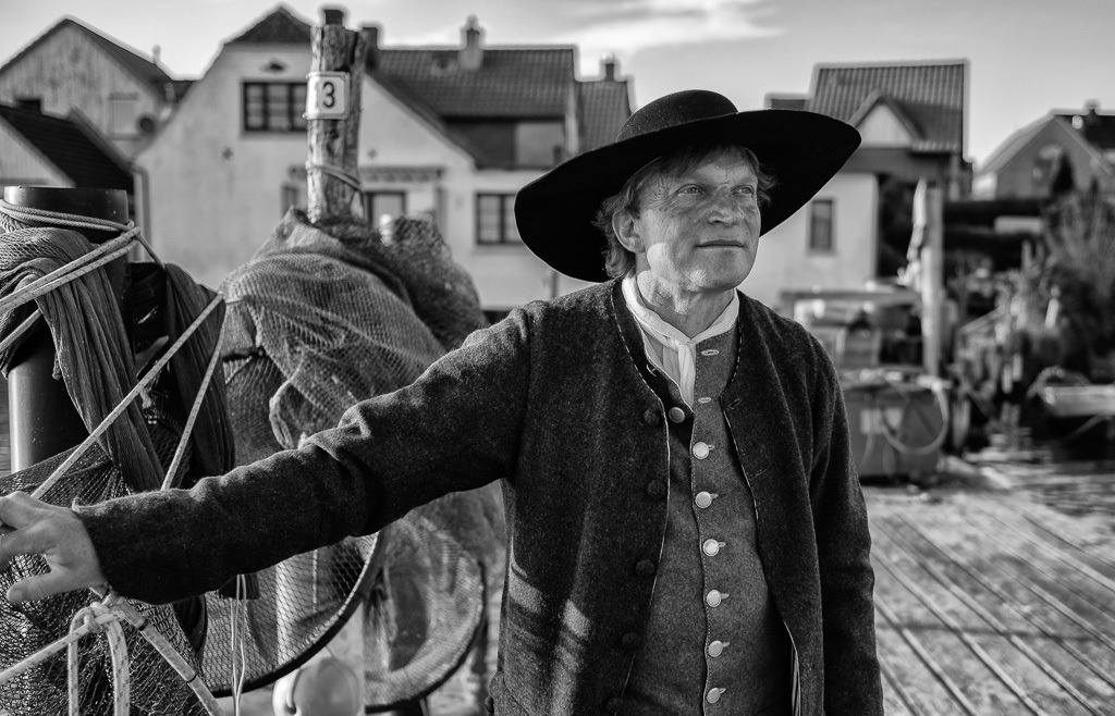 fischer-2020-086 | Jörg Nadler ist Mitglied der Holmer Fischerzunft in Schleswig. Er nennt sich "historischer Fischer", weil er neben seinem Hauptberuf das Fischerhandwerk früherer Epochen als Darsteller u. a. bei museumspädagogischen Veranstaltungen lebendig vermittelt. Hier ist er als Fischer der Barockzeit auf seiner Brücke am Ufer des Holms zu sehen. - Realisiert mit Pictrs.com