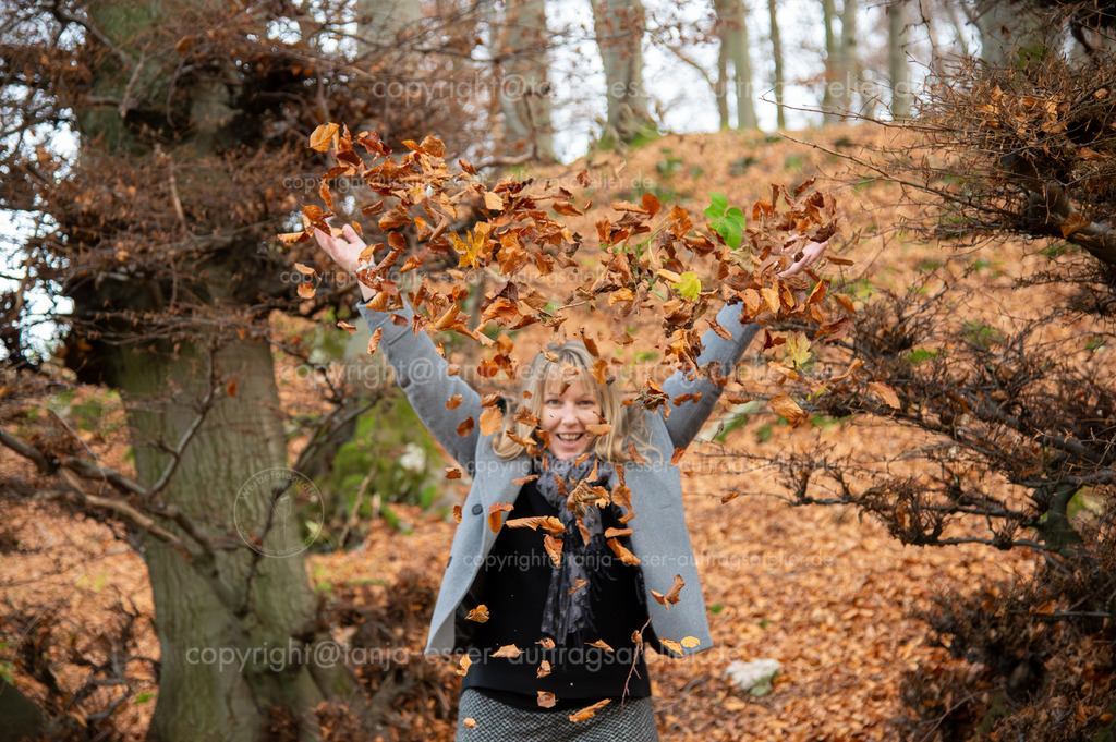 Eine 40 jährige blonde Frau in grauer eleganter Kleidung wirft voller Lebensfreude Herbstlaub in die Höhe. Im Hintergrund ist unscharf ein herbstlicher Laubwald zu sehen. | Eine 40 jährige blonde Frau in grauer eleganter Kleidung wirft voller Lebensfreude Herbstlaub in die Höhe. Im Hintergrund ist unscharf ein herbstlicher Laubwald zu sehen.