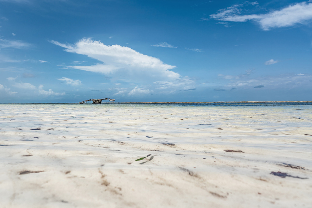 Paradiesischer Strand | Kenia | Premium-Wandbild von justsomewhere.com: Ein endloser, schneeweißer Sandstrand an der Küste Kenias geht bei Ebbe in flaches, kristallklares Wasser über, unter einem weiten, strahlend blauen Himmel mit dramatischen Wolken. - Realisiert mit Pictrs.com