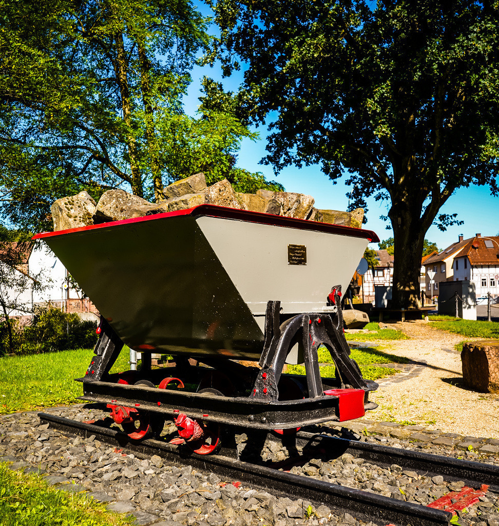 Lore - Rinderbügen | Eine Lore mit roten Akzenten steht in sinnbildlich für die Bergbauvergangenheit in der Region. Auch in Rinderbügen zeigt der Sommer an diesem Wochenende ein letztes Mal was in im steckt. In der sengenden Mittagshitze entfalten die Exponate in der Dorfmitte eine eigene Atmosphäre und die Kunstwerke aus Eisen und Stein werden durch die kontrastspendenden Schatten zum Leben erweckt. Als Besucher spürt man die Liebe und Mühe, die in die Gestaltung und Erhaltung des Platzes fließen. Vielleicht dient dieser Ort als Inspiration für die neuen Dorfmitten in Gedern und Hirzenhain? - Realisiert mit Pictrs.com