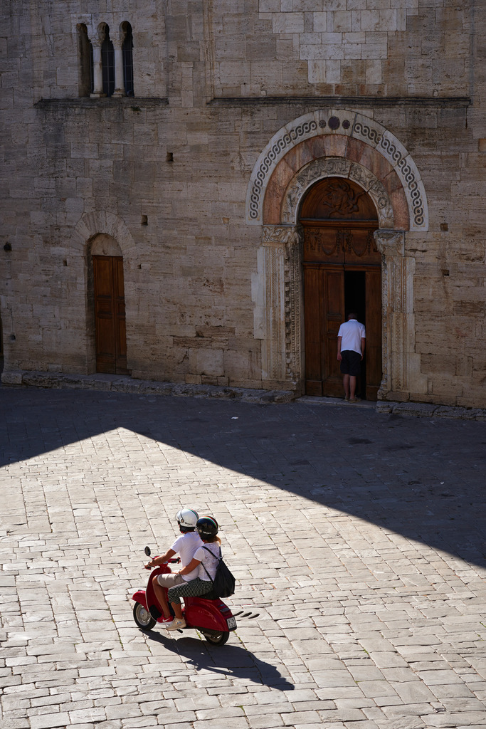 Junges Paar auf einer grauen Vespa | Bevagna, Italien - August 28, 2016: Junges Paar auf einer grauen Vespa fährt über den Dorfplatz. - Realisiert mit Pictrs.com