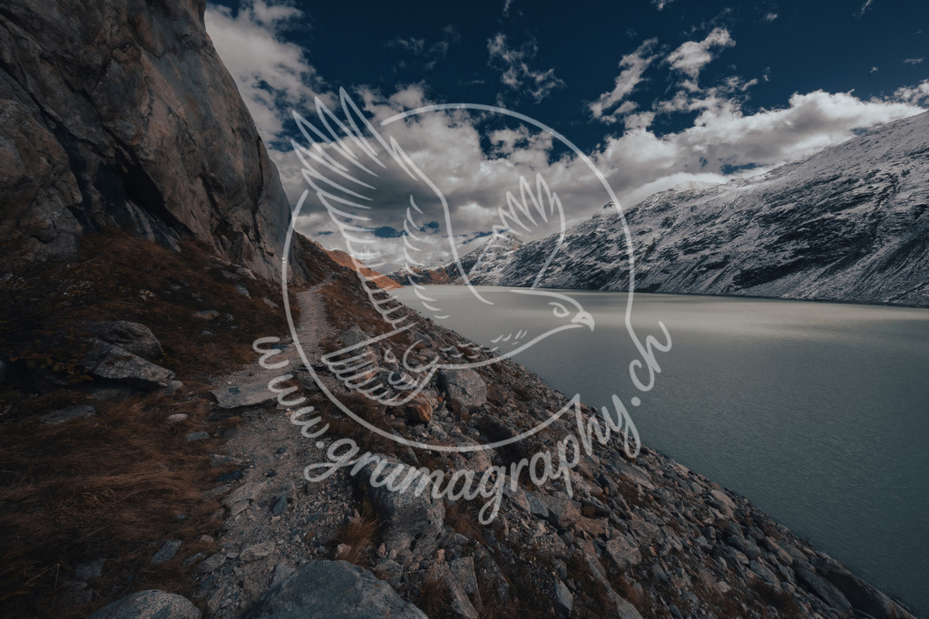 along the silent shore - switzerland | Diese Aufnahme zeigt einen schmalen Weg entlang eines hochalpinen Stausees in den Schweizer Alpen. Steile Felsflanken, ruhiges Wasser und ein zurückhaltendes Wolkenbild prägen die Szene. Gedämpfte Farben, klare Flächen und eine reduzierte Bildsprache betonen die stille Atmosphäre und die Weite der Landschaft. Die Fotografie wurde bewusst ohne Inszenierung aufgenommen und im charakteristischen Grumagraphy-Style entwickelt, mit Fokus auf Ruhe, Zeitlosigkeit und natürlicher Balance.