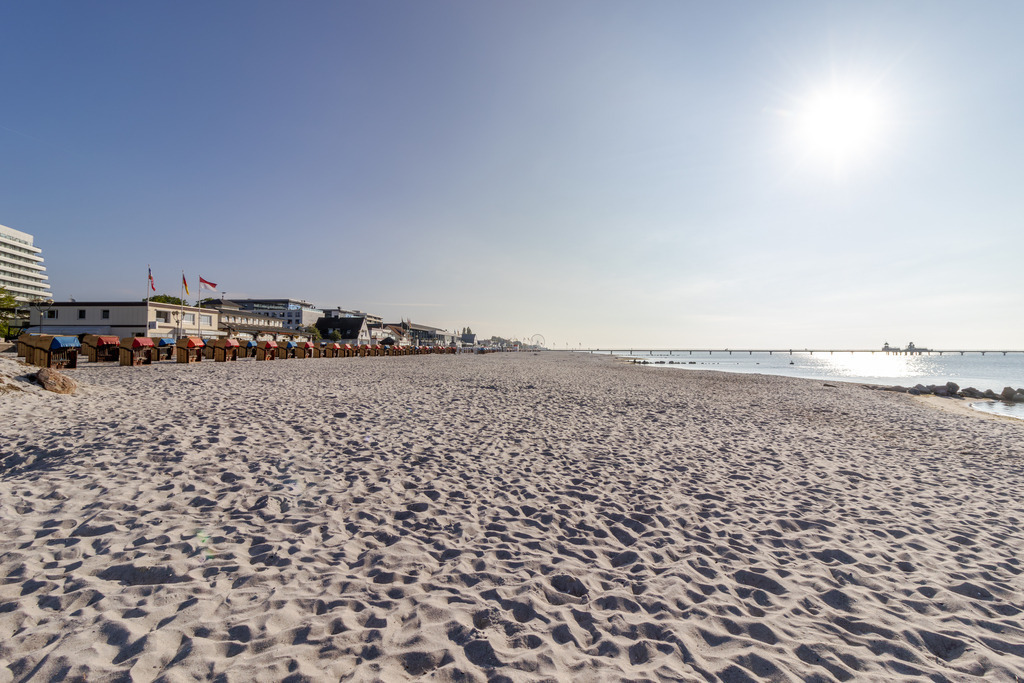 Wandbild: Grömitz erwacht  | Dieses Wandbild zeigt eine weitläufige Strandszene in Grömitz an einem sonnigen Morgen. Der helle Sand erstreckt sich bis zum Horizont, durchzogen von Fußspuren, die von früher Aktivität zeugen. Links im Bild stehen zahlreiche Strandkörbe mit Sonnendächern in geordneten Reihen – bereit für einen Tag voller Ruhe und Erholung. Dahinter erheben sich mehrere Gebäude, darunter Hotels und Strandbetriebe, von denen einige mit wehenden Fahnen geschmückt sind. In der Ferne zieht sich die Seebrücke ins Wasser, ein markantes Element der Grömitzer Küstenarchitektur. Der Himmel ist klar und strahlt in hellem Blau, das Licht fällt weich über die Szene und unterstreicht die ruhige, strukturierte Atmosphäre. Dieses Motiv eignet sich ideal als Wandbild für maritime Wohnkonzepte – ob als Leinwandbild, Acrylglasbild, Alu-Dibond FineArt Print oder als Akustikbild. Ein stilvoller Akzent für Wohnzimmer, Büro oder Ferienwohnung. - Realisiert mit Pictrs.com