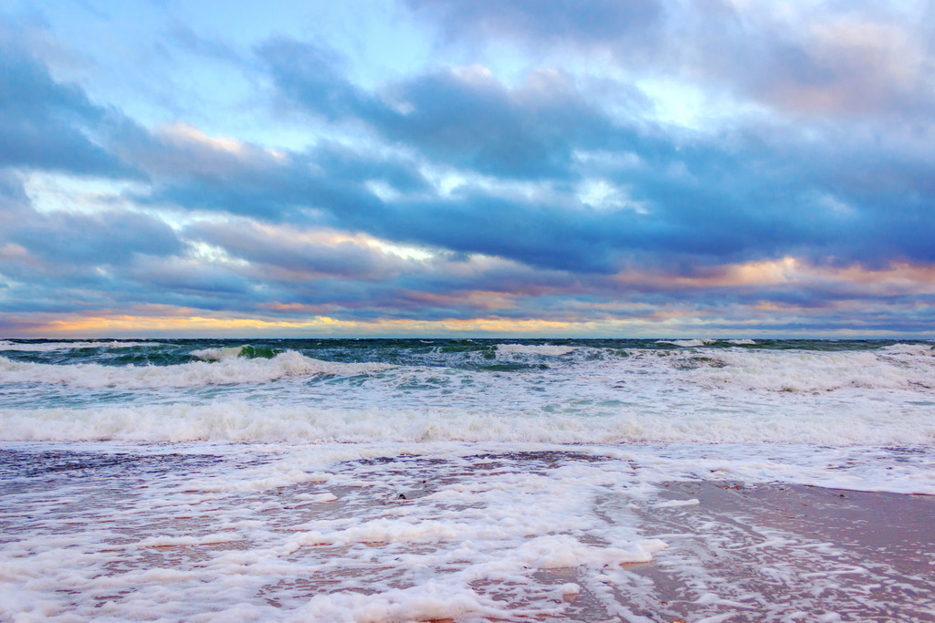 Wandbild: Morgenstimmung am Meer | Dieses Wandbild im Querformat zeigt die stürmische Ostsee in einer schönen Morgenstimmung. Im Vordergrund ist auf dem Strand der Schaum der Gischt zu sehen. Auf dem Meer befinden sich Wellen mit schönen Schaumkronen. Das Licht der aufgehenden Sonne bringt verschiedene warme Farbtöne ins Wandbild.  - Realisiert mit Pictrs.com
