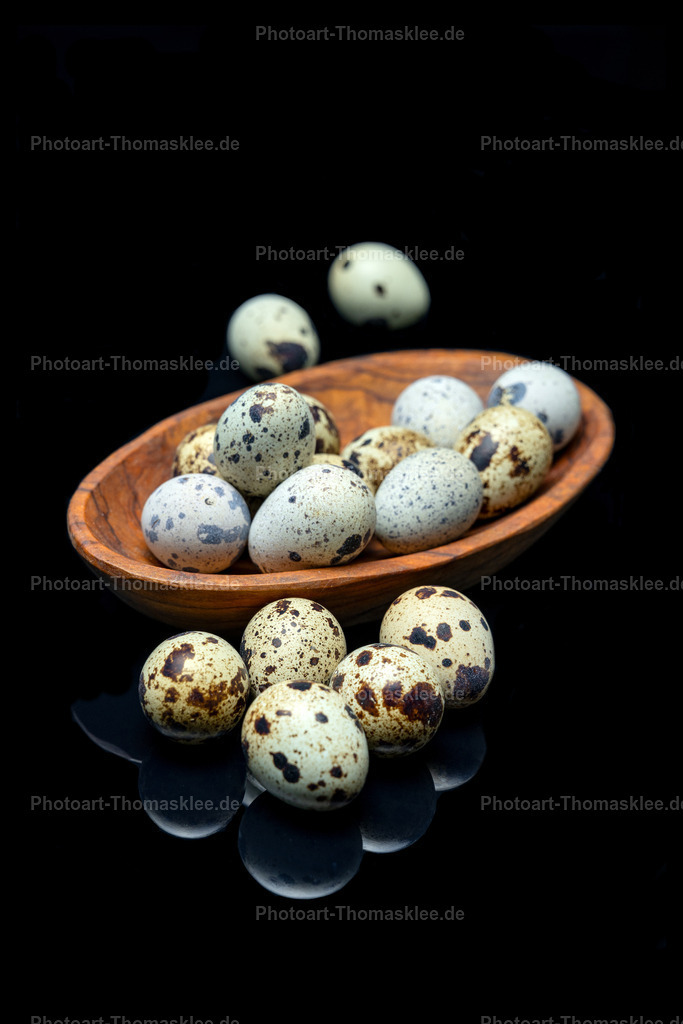 Wachteleier in Olivenholzschale auf schwarzem Glas | Diese Fine-Art-Fotografie zeigt frische Wachteleier in einer handgefertigten Olivenholzschale vor tiefschwarzen Hintergrund. Die gesprenkelten Eier ruhen auf einer schwarzen Glasplatte, die faszinierende Reflexionen erzeugt. Einzelne Wachteleier liegen außerhalb der Schale und verstärken die natürliche Komposition. Die warmen Brauntöne des Olivenholzes kontrastieren perfekt mit der kühlen Eleganz des Glases. Dramatische Beleuchtung hebt die feinen Strukturen der Eierschalen hervor und schafft eine zeitlose Stillleben-Atmosphäre. - Realisiert mit Pictrs.com