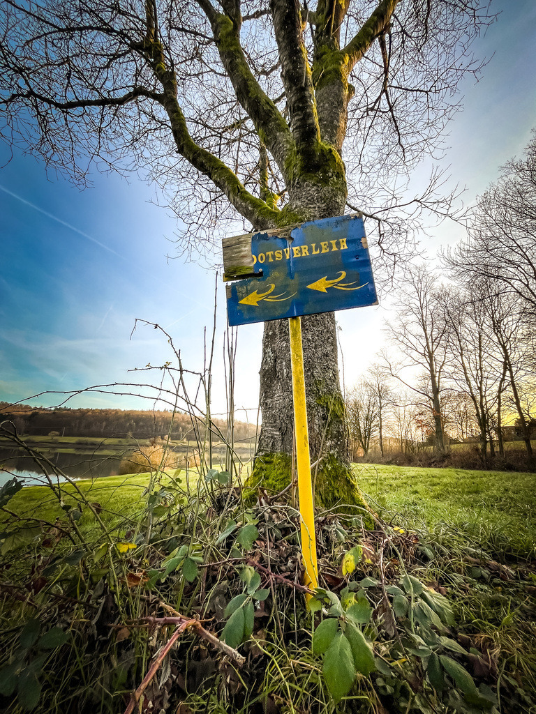 Ootsverleih | IMG_0037:Der Bootsverleih am Nidda-Stausee sucht derzeit nach einem neuen Anfangsbuchstaben.Der Nidda-Stausee schafft im Dezember eine Atmosphäre der Gegensätze. Während die Bäume bereits ihr Blätterkleid für den Winter abgelegt haben, wirkt das Gras grün und üppig. Während am Horizont die heimischen Wälder zu sehen sind, scheint das unmittelbare Seeufer fremd, steinig und kahl. Zusammen schaffen diese Eindrücke eine einzigartige Kulisse, die auch abseits der Badesaison einen Besuch wert ist. - Realisiert mit Pictrs.com