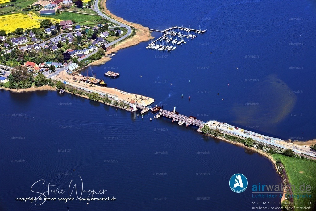 Luftbild Ostseefjord Schlei, Boren-Lindaunis Schleibrücke, Campingplatz Lindaunis, Sportboothafen Lindaunis | Die Schleibrücke Lindaunis, eine historische Rollklappbrücke über den Meeresarm Schlei, hat eine bedeutende Geschichte im 20. Jahrhundert. Ihre Vorgängerin war eine Drehbrücke, die von 1881 bis 1926 an derselben Stelle existierte und die Eisenbahnstrecke Kiel–Flensburg über die Schlei verband. Aufgrund der zu geringen Schifffahrtsöffnung wurde die Drehbrücke durch eine neue Klappbrücke ersetzt, die am 17. Juli 1927 in Betrieb ging. Die neue Brücke wurde aus Teilen der abgebauten Drehbrücke Taterpfahl gebaut, die 1920 durch eine moderne Hochbrücke ersetzt wurde. Die Schleibrücke Lindaunis verbindet die Halbinseln Schwansen und Angeln und wurde 1997 unter Denkmalschutz gestellt. Sie wurde ursprünglich mit einem Kettenantrieb betrieben, ab 1975 erfolgte die Bewegung hydraulisch. Die Brücke ist seit 2021 für Eisenbahnen und Kraftfahrzeuge gesperrt und kann nur noch zu Fuß überquert werden, da die alte Brücke abgebaut und durch einen Neubau ersetzt wird.