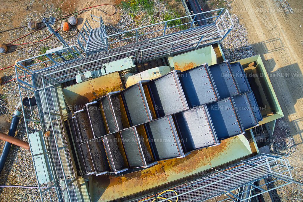 hülskens_2_0032 | Senkrechter Blick aus der Drohne auf das Schöpfrad im Kieswerk Rothensee. Die Kamera befindet sich dabei direkt über der Anlage und zeigt ausschließlich die markant angeordneten Schaufeln aus unmittelbarer Nähe.Durch die senkrechte Perspektive entsteht ein ungewöhnliches, fast grafisches Motiv. Die regelmäßige Anordnung der Schaufeln, die Stahlkonstruktion und die geometrischen Formen lassen die technische Anlage beinahe abstrakt wirken.Damit bildet diese Aufnahme den Abschluss der kleinen Kieswerk-Serie. Nach dem großräumigen Blick auf das Abbaugebiet und das Werk, den näheren Drohnenaufnahmen und den klassischen Detailaufnahmen des Schöpfrades endet die Serie mit einer bewusst ungewöhnlichen Perspektive aus der Drohne.Ein technisches Detail wird zum eigenständigen Fotomotiv – genau die Art von Aufnahme, die zeigt, wie vielseitig professionelle Industriedokumentation sein kann. - Realisiert mit Pictrs.com