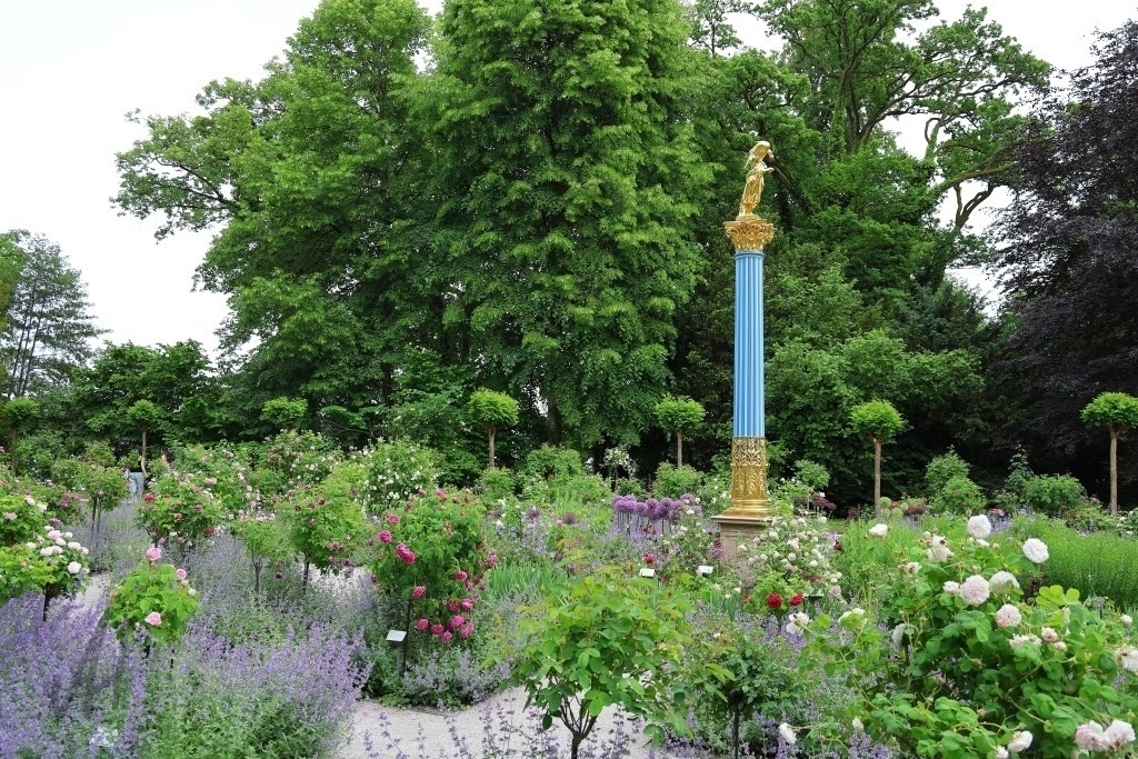 Roseninsel im Starnberger See | Im idyllischen Rosengarten auf der Roseninsel im Starnberger See inmitten sorgfältig angelegter Beete mit blühenden Rosen erhebt sich der goldene Engel als strahlender Blickfang der Anlage. Seine vergoldete Oberfläche reflektiert das Sonnenlicht und setzt einen leuchtenden Akzent zwischen den Farben der Blumen und dem satten Grün der Gartenanlage. - Realisiert mit Pictrs.com