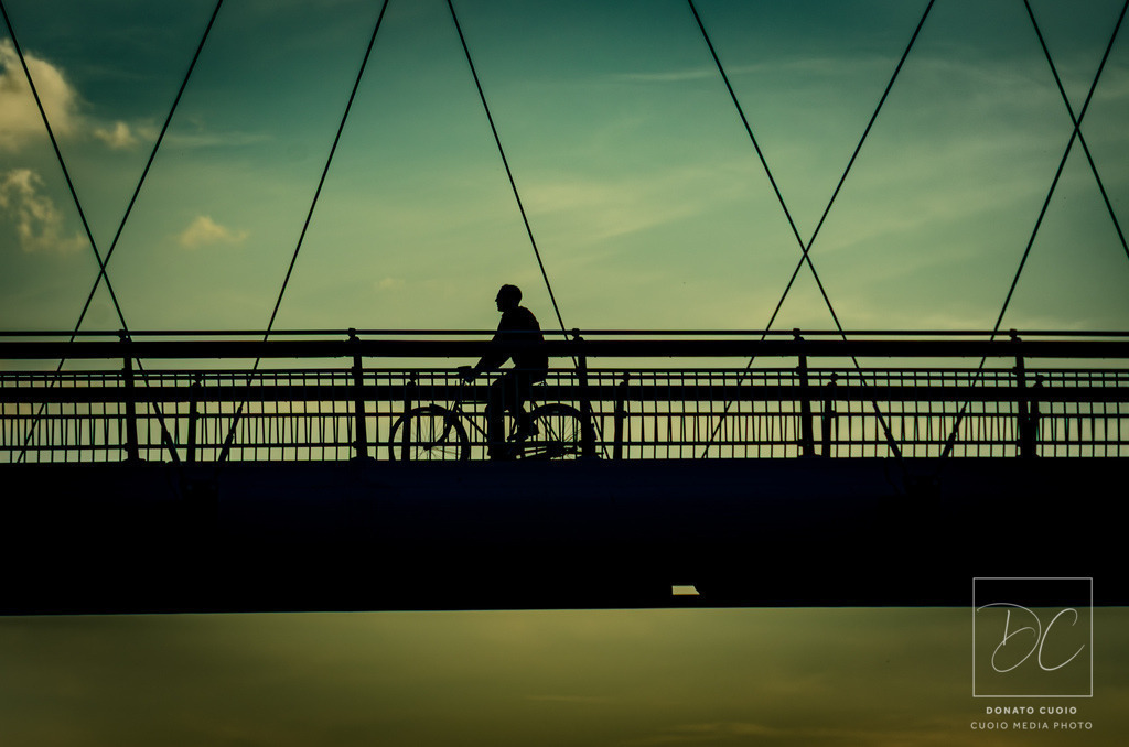 Silhouettenfahrt auf dem Holbeinsteg bei Sonnenuntergang | Ein Radfahrer überquert den Holbeinsteg in Frankfurt bei Sonnenuntergang. Die Silhouette hebt sich eindrucksvoll vor dem warmen Himmel ab. Die Seilstruktur der Brücke erzeugt eine geometrische Dynamik, während die Abendstimmung eine ruhige Atmosphäre schafft. Ein Moment von Bewegung und Stille zugleich. - Realisiert mit Pictrs.com