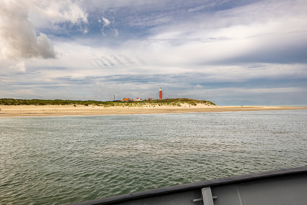Blick von See auf den Leuchtturm Eierland und die Nordküste von Texel | Panoramaaufnahme vom Wasser aus auf die Nordspitze der holländischen Insel Texel. Im Zentrum der Küstenlinie erhebt sich der rote Leuchtturm Eierland hinter der weitläufigen Dünen- und Strandlandschaft. Die Aufnahme vermittelt die Weite der Nordsee bei wechselnder Bewölkung und eignet sich ideal für redaktionelle Beiträge zu Inseltourismus, Seefahrt, Urlaub in den Niederlanden und Küstenschutz. - Realisiert mit Pictrs.com