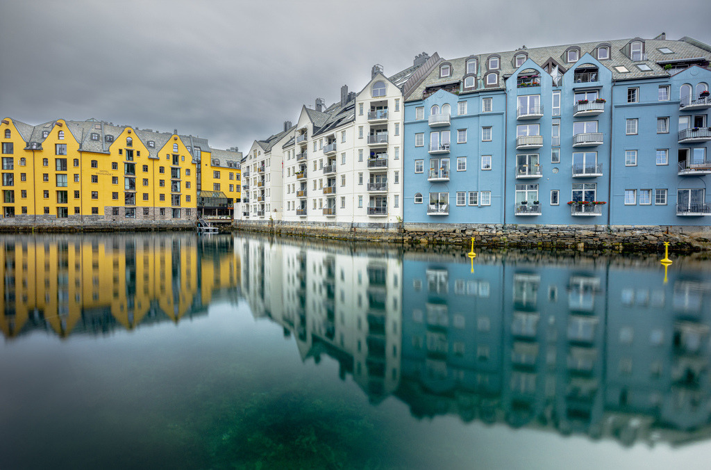Spiegelnde Häuser in der Innenstand von Ålesund---Norwegen | Die Abbildung zeigt den Brosundet-Kanal in Ålesund, Norwegen, bekannt für seine einzigartige Jugendstil-Architektur. Die Stadt wurde nach einem verheerenden Brand im Jahr 1904 fast vollständig im Jugendstil wiederaufgebaut. Die farbenfrohen Gebäude spiegeln sich im Wasser des Kanals, der die Inseln Aspøy und Nørvøy voneinander trennt. Ålesund liegt an der Westküste Norwegens und ist ein wichtiger Anlaufpunkt für Kreuzfahrten und Hurtigruten-Schiffe.  - Realisiert mit Pictrs.com