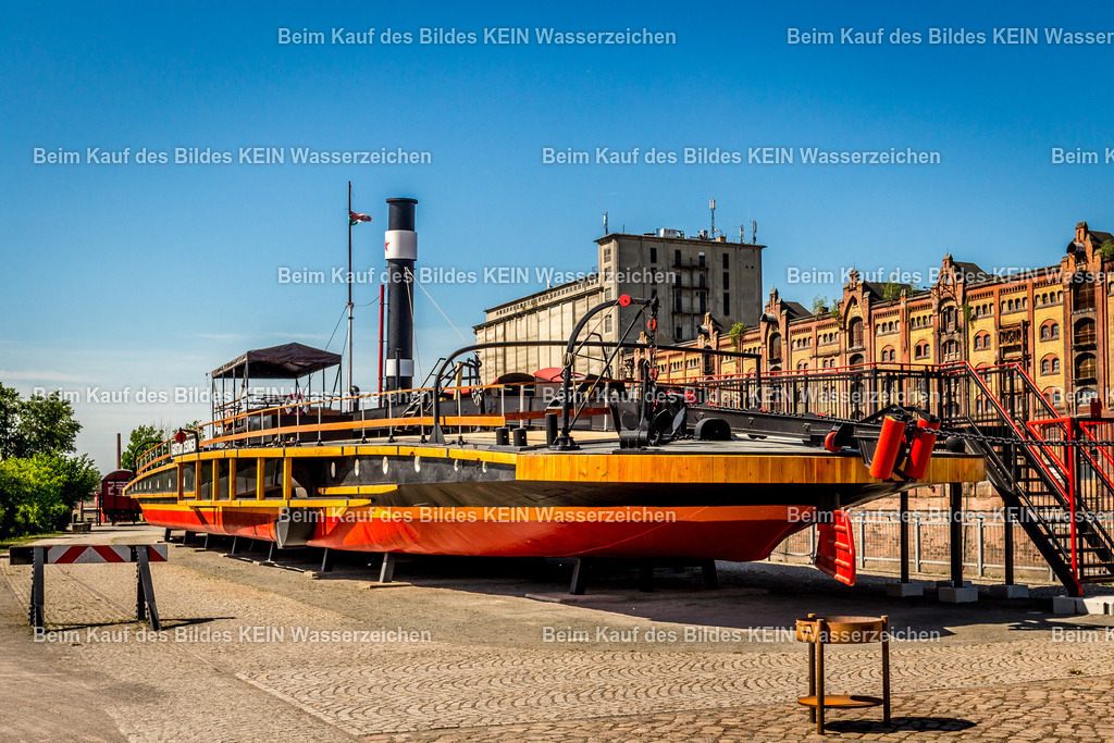 Magdeburg Gustav Zeuner-9463 | Das Foto zeigt den historischen Kettenschleppdampfer „Gustav Zeuner“ im Wissenschaftshafen Magdeburg aus einer bodennahen Perspektive. Die genietete Stahlkonstruktion, der hohe Schornstein und die markanten Aufbauten vermitteln einen eindrucksvollen Eindruck der technischen Entwicklung der Elbschifffahrt im späten 19. Jahrhundert. Die niedrige Kameraposition betont die Länge und die robuste Bauweise des Schiffes.Die „Gustav Zeuner“ wurde 1894 auf der Schiffswerft Übigau bei Dresden gebaut und war der erste Kettenschleppdampfer der zweiten Generation auf der Elbe. Das Schiff ist 55,47 Meter lang, 10,34 Meter breit und wurde von zwei Verbunddampfmaschinen mit einer Gesamtleistung von 180 PS angetrieben. Über ein Greifrad griff der Dampfer die auf dem Flussgrund verlegte Schleppkette und konnte so schwere Lastkähne stromaufwärts bewegen – ein für seine Zeit äußerst effizientes Transportsystem. Heute ist die „Gustav Zeuner“ der einzige nahezu vollständig erhaltene Kettenschleppdampfer der Elbe und ein bedeutendes technisches Denkmal der Binnenschifffahrt. Die Aufnahme dokumentiert eindrucksvoll die Ingenieurtechnik einer längst vergangenen Epoche der Schifffahrt. Im Wissenschaftshafen Magdeburg erinnert die „Gustav Zeuner“ als Museumsschiff an die Entwicklung des Gütertransports auf der Elbe und an eine Technik, die den Binnenschiffsverkehr über Jahrzehnte entscheidend geprägt hat. - Realisiert mit Pictrs.com