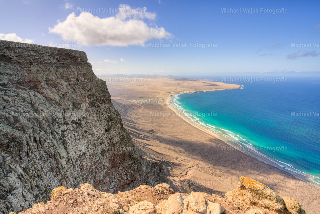 Blick vom Mirador de El Risco de Famara auf Lanzarote | Ein Blick vom Mirador de El Risco de Famara in Richtung der mächtigen Famara‑Klippe und des endlosen Famara‑Strands hat etwas Erhabenes. Die Steilwand zieht sich wie ein gewaltiger Rücken aus Vulkangestein entlang der Küste, während unten der helle Sandstreifen sanft vom Atlantik umspült wird. An einem klaren Tag verschmelzen Farben und Formen zu einer beeindruckenden Szenerie: das tiefe Blau des Meeres, die goldenen Dünen, die schroffe Felswand – ein Panorama, das Ruhe ausstrahlt und gleichzeitig die wilde Energie Lanzarotes spürbar macht. - Realisiert mit Pictrs.com