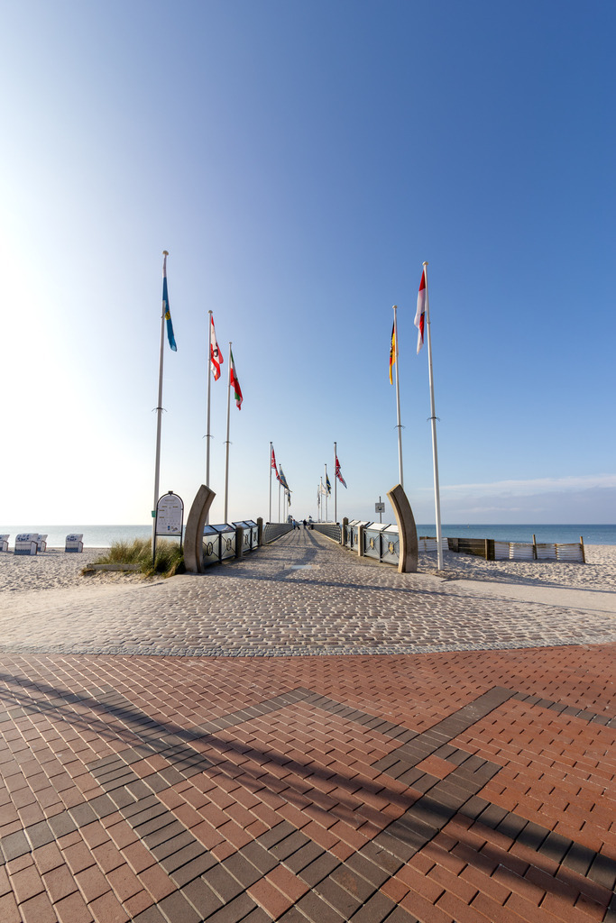 Wandbild: Seebrücke mit Weitblick in Grömitz | Dieses Wandbild zeigt den Beginn der Seebrücke von Grömitz an einem sonnigen Morgen. Die gepflasterte Promenade ist mit roten und braunen Steinen in geometrischem Muster ausgelegt und führt geradlinig auf das Meer hinaus. Entlang der Brücke stehen hohe Fahnenmasten. Links und rechts am Brückeneingang rahmen geschwungene Metallstrukturen den Zugang und setzen gestalterische Akzente. Im Hintergrund sind Strandkörbe auf dem Sand verteilt, dahinter das ruhige Meer unter einem wolkenlosen Himmel. Die Komposition wirkt symmetrisch und strukturiert, zugleich offen und einladend – ein Moment der Klarheit und Küstenruhe. Dieses Motiv eignet sich ideal als Wandbild für maritime Wohnkonzepte – ob als Leinwandbild, Acrylglasbild, Alu-Dibond FineArt Print oder als Akustikbild. Ein stilvoller Akzent für Wohnzimmer, Büro oder Ferienwohnung. - Realisiert mit Pictrs.com
