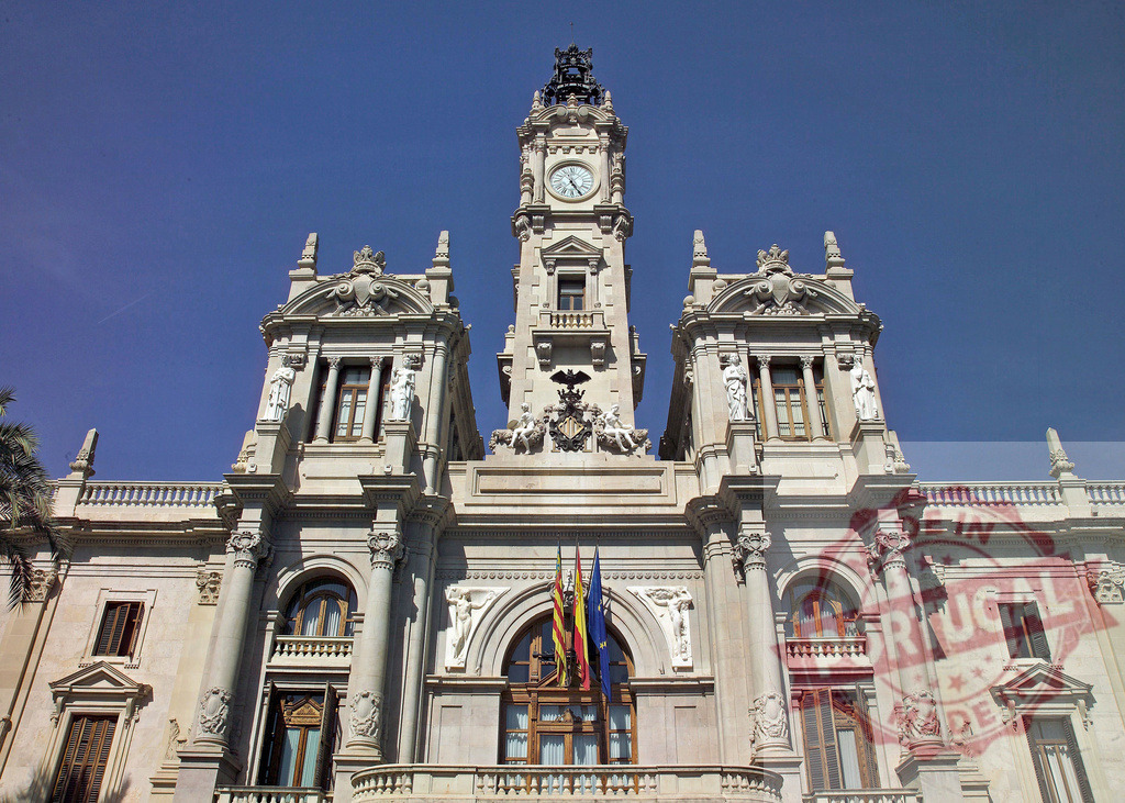 Valencia City Hall exhibiting intricate architecture and Spanish flags waving proudly | Majestic Valencia City Hall stands tall, showcasing elaborate architecture, sculptures, clock tower, and Spanish flags against a vibrant blue sky - Realized with Pictrs.com