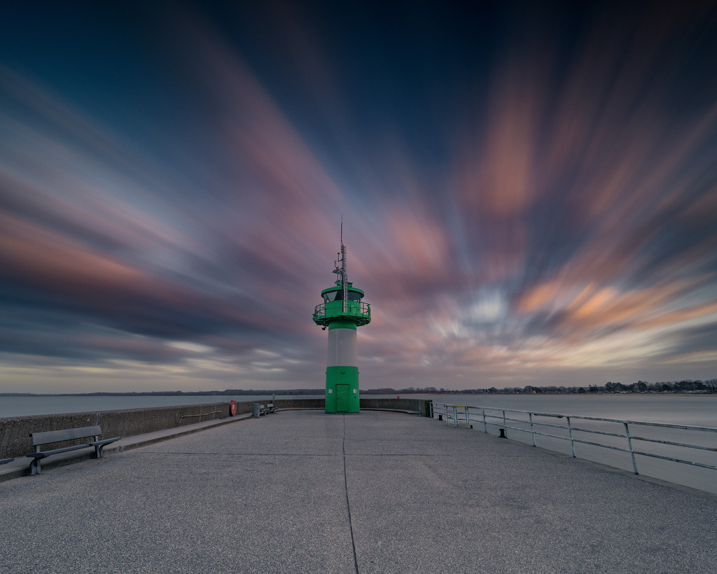 Grünes Molenfeuer unter ziehenden Wolken | Das grüne Molenfeuer steht am Ende der Mole unter einem dramatisch gefärbten Himmel. Die Langzeitbelichtung zieht die Wolken in kraftvollen Linien über das Motiv und betont die symmetrische Perspektive. - Realisiert mit Pictrs.com