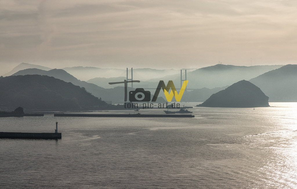 Die Bucht von Nagasaki im früh morgendlichen Dunst---Japan | Das Bild zeigt die Bucht von Nagasaki in Japan, mit einem deutlichen Blick auf die künstliche Insel Dejima. Diese Insel spielte eine zentrale Rolle in der japanischen Geschichte. Hier sind die wichtigsten Details zu Dejima:Historische Bedeutung: Dejima war während der Edo-Zeit (1615–1868) der einzige Ort, an dem westliche Händler, insbesondere die Niederländer, Handel mit Japan treiben durften. Isolation: Die fächerförmige, künstlich aufgeschüttete Insel wurde 1636 erbaut, um ausländische Händler vom japanischen Festland zu trennen und die Ausbreitung des Christentums zu kontrollieren.  - Realisiert mit Pictrs.com