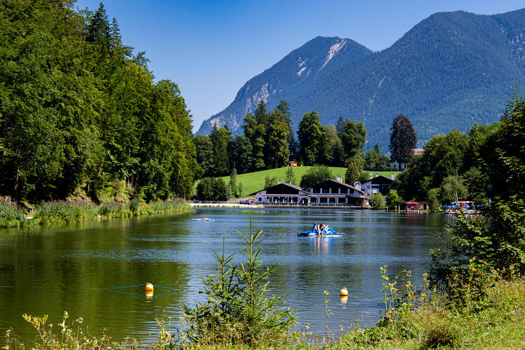 Idyllisches Badevergnügen: Sommertag am Riessersee | Ein traumhafter Sommertag wie aus dem Bilderbuch: Auf der ruhigen, grün schimmernden Wasseroberfläche des historischen Riessersees bei Garmisch-Partenkirchen genießen Ausflügler im Tretboot und auf Stand-Up-Paddle-Boards die erfrischende Auszeit. Am Ufer fügt sich das bekannte Seehaus malerisch in die Natur ein, während im Hintergrund die bewaldeten Hänge und imposanten Gipfel des Wettersteingebirges emporragen. Eine herzerwärmende Aufnahme, die bayerische Gemütlichkeit, Sommerglück und pure Erholung vor alpiner Kulisse ausstrahlt. - Realisiert mit Pictrs.com