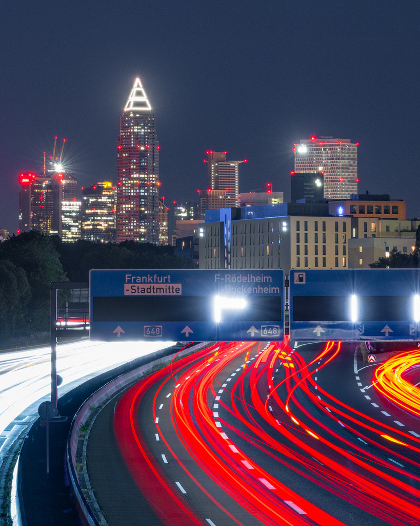 Lichtspuren vor der Frankfurter Skyline | Rote und weiße Lichtspuren führen über die Autobahn direkt auf die nächtliche Skyline von Frankfurt zu. Die Langzeitbelichtung verbindet urbanen Verkehr und Hochhausarchitektur zu einer dynamischen Stadtansicht. - Realisiert mit Pictrs.com
