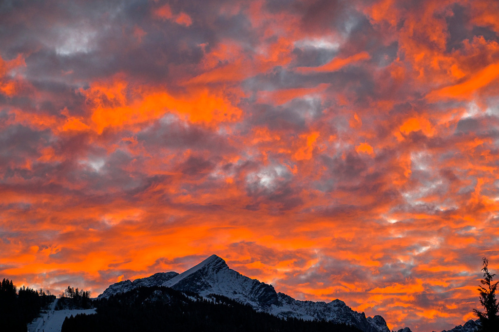 Brennender Himmel: Das dramatische Glühen über der Alpspitze | Ein Anblick von überwältigender Intensität und wilder, emotionaler Kraft: Wie ein gewaltiges Himmelsfeuer lodert ein dramatischer Wolkenhimmel in tiefem Orangerot und brennendem Gelb auf. Die zerrissenen, dunklen Wolkenformationen verstärken das feurige Leuchten, das eine fast mystische Energie verströmt. Ruhig, erhaben und unbeeindruckt von diesem glühenden Schauspiel ragt die markante, schneebedeckte Pyramide der Alpspitze am Horizont empor. Im tiefen Schatten des Vordergrunds zeichnen sich die dunklen Silhouetten der Wälder und Skipisten ab. Diese Aufnahme fängt ein faszinierendes Naturschauspiel voller Kontraste ein – ein Moment, der die majestätische Urgewalt der Bergwelt spürbar macht. - Realisiert mit Pictrs.com
