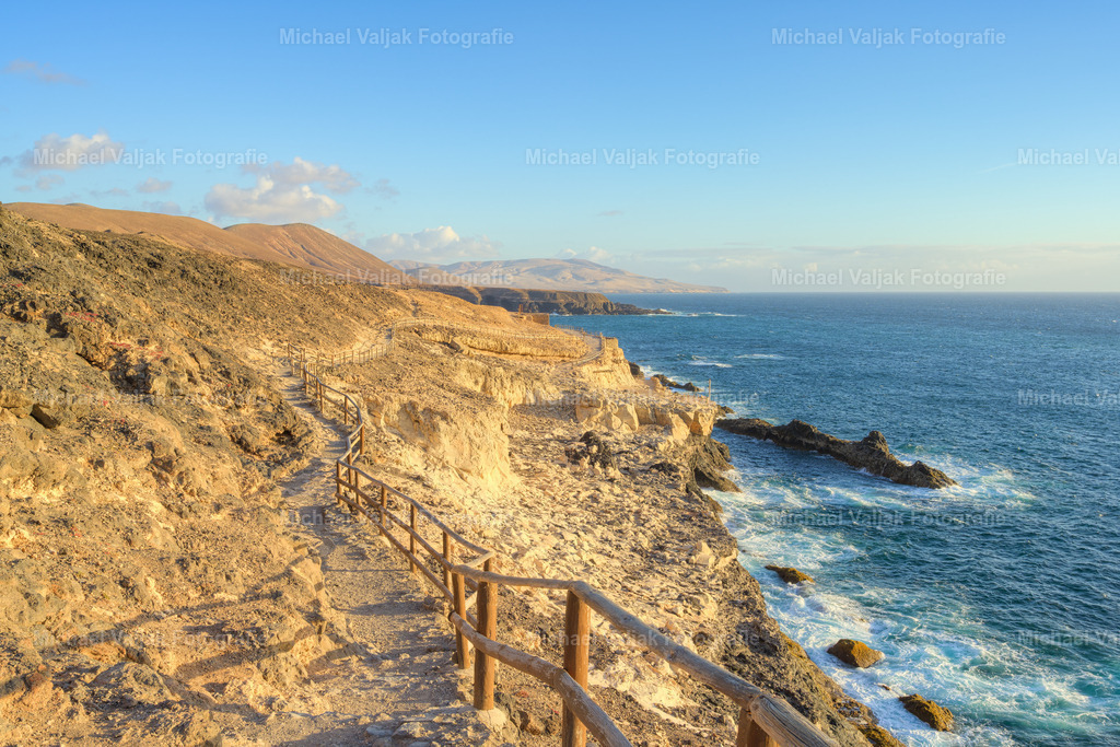 Küstenweg in Ajuy auf Fuerteventura | Der Weg zu den Höhlen von Ajuy verläuft über hellen, sonnengebleichten Fels, der sich deutlich von den dunklen Klippen der Westküste absetzt. Der Pfad folgt der natürlichen Form der Küste und öffnet immer wieder den Blick auf den tiefblauen Atlantik, der unterhalb gegen die Felsen bricht. Das Licht betont die warmen Töne des Gesteins und die klare Struktur des Weges. Ein Moment, in dem die helle Felslandschaft und die Kraft des Meeres eine eindrucksvolle Kombination bilden. - Realisiert mit Pictrs.com
