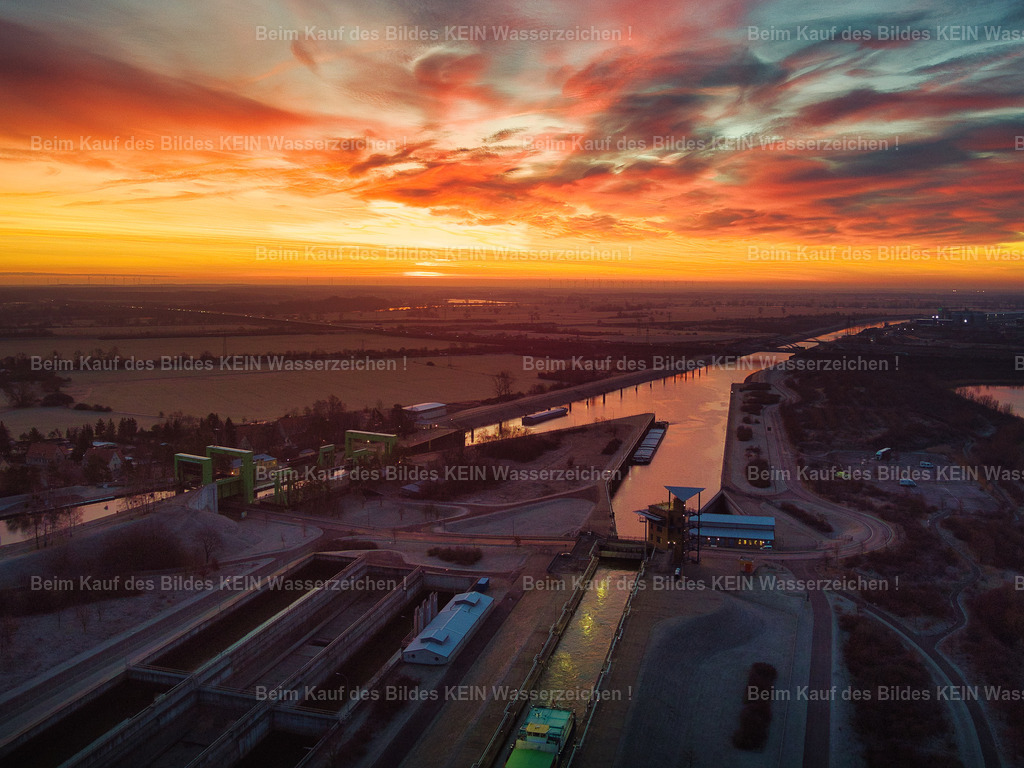 Magdeburg_Sonnenaufgang_Schleuse_Rothensee_roter_Himmel_0034 | Dieses Luftbild zeigt zwei der bedeutendsten Wasserbauwerke Magdeburgs in den frühen Morgenstunden eines farbenprächtigen Sonnenaufgangs. Im Vordergrund erstreckt sich die moderne Doppelsparschleuse Rothensee, während dahinter das historische Schiffshebewerk Rothensee zu erkennen ist. Gemeinsam dokumentieren beide Bauwerke die Entwicklung der Wasserbautechnik über mehrere Generationen hinweg.Die Aufnahme wird von einer außergewöhnlichen Lichtstimmung geprägt. Intensive Rot-, Orange- und Blautöne am Himmel spiegeln sich in den Wasserflächen des Hafenbeckens wider und verleihen der Szene eine eindrucksvolle Atmosphäre. Die ersten Sonnenstrahlen des Tages setzen die technischen Bauwerke wirkungsvoll in Szene und betonen die klaren Linien der Schleusen- und Hafenanlagen.Aus der Vogelperspektive wird die besondere Bedeutung dieses Bereichs für die Binnenschifffahrt sichtbar. Hier treffen moderne Infrastruktur, historische Ingenieurskunst und die weitläufigen Hafenanlagen aufeinander. Das Luftbild zeigt eindrucksvoll die Verbindung von Technik, Wasser und Landschaft und vermittelt einen faszinierenden Blick auf einen der wichtigsten Verkehrsknotenpunkte der deutschen Wasserstraßen. - Realisiert mit Pictrs.com