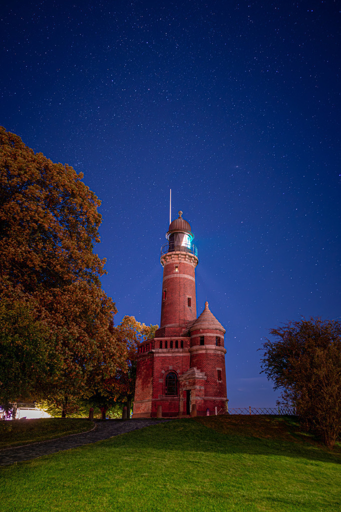 Leuchturm Kiel Holtenau mit Sternenhimmel  | Eine beeindruckende Nachtaufnahme des Leuchtturms Holtenau. Das Licht des Leuchtturms erzeugt einen hellen, sichtbaren Lichtstrahl, der sich in der Dunkelheit des Nachthimmels abzeichnet. Die Struktur des Turms ist im Kontrast zur Dunkelheit des Himmels und des umliegenden Geländes gut erkennbar. Das Leuchtfeuer des Holtenau-Leuchtturms weist Schiffen den sicheren Weg durch die Nacht und ist ein Zeichen der Hoffnung und des Schutzes in einer dunklen Umgebung. Es ist ein wundervolles Beispiel für die maritime Architektur und die Rolle, die Leuchttürme in der Geschichte der Seefahrt spielen.