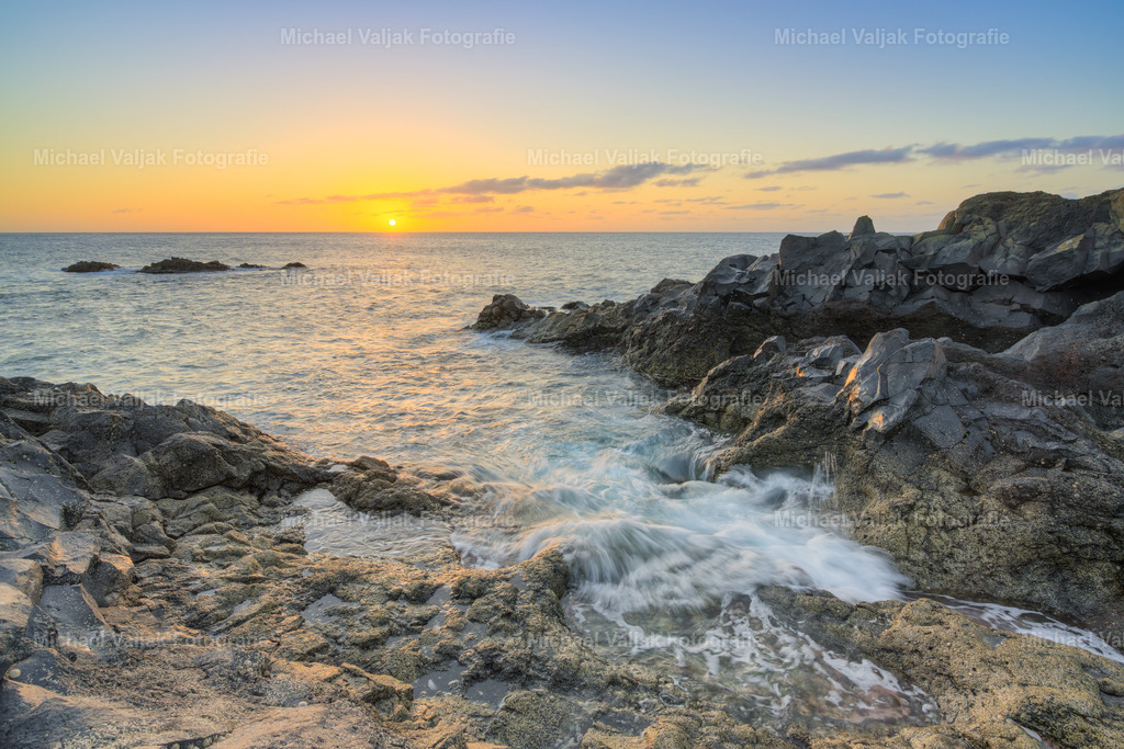 Sonnenuntergang an der rauen Westküste von Lanzarote | An der Westküste Lanzarotes, unweit der tosenden Felsformationen von Los Hervideros, zeigt der Sonnenuntergang seine kraftvollste Seite. Die Sonne sinkt langsam hinter den Atlantik, färbt den Horizont in tiefe Gold‑ und Kupfertöne und lässt die schwarzen Lavaklippen für einen Moment warm aufglühen. Das Foto hält diesen Übergang fest – ein stiller Augenblick in einer Landschaft, die sonst von Bewegung und Energie geprägt ist. - Realisiert mit Pictrs.com
