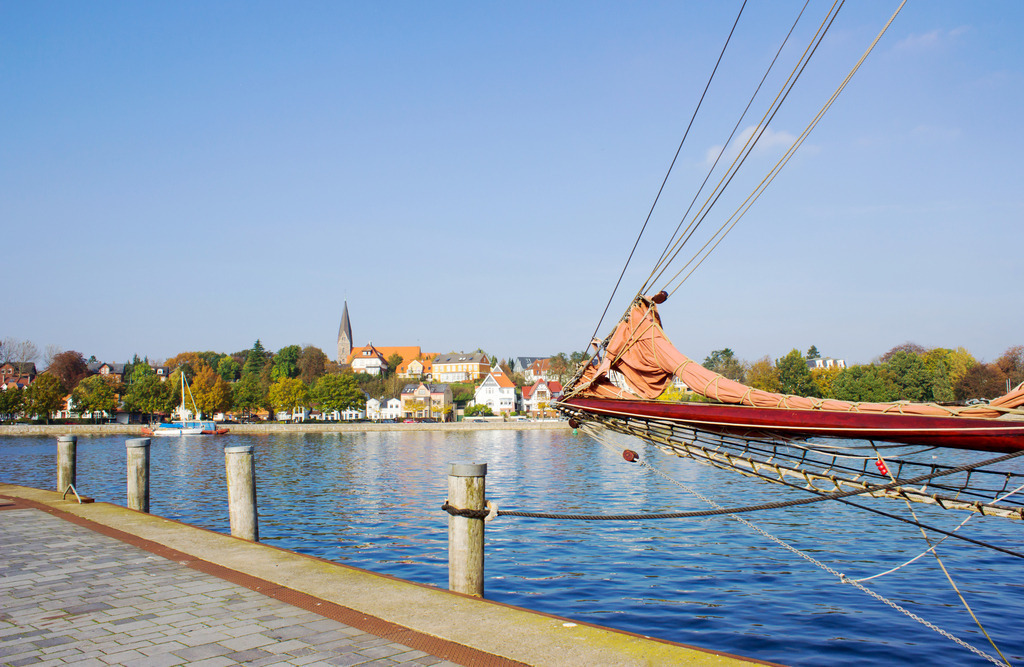 Wandbild: Hafen in Eckernförde mit Blick zur Kirche in Borby | Dieses Wandbild im Querformat zeigt den Hafen in Eckernförde im Herbst. Von der rechten Seite ragt die Takelage eines Traditionsseglers ins Bild. In der Ferne kann man auf der anderen Seite des Hafens die  Kirche in Borby erkennen. Der blaue Himmel ist fast wolkenlos. - Realisiert mit Pictrs.com
