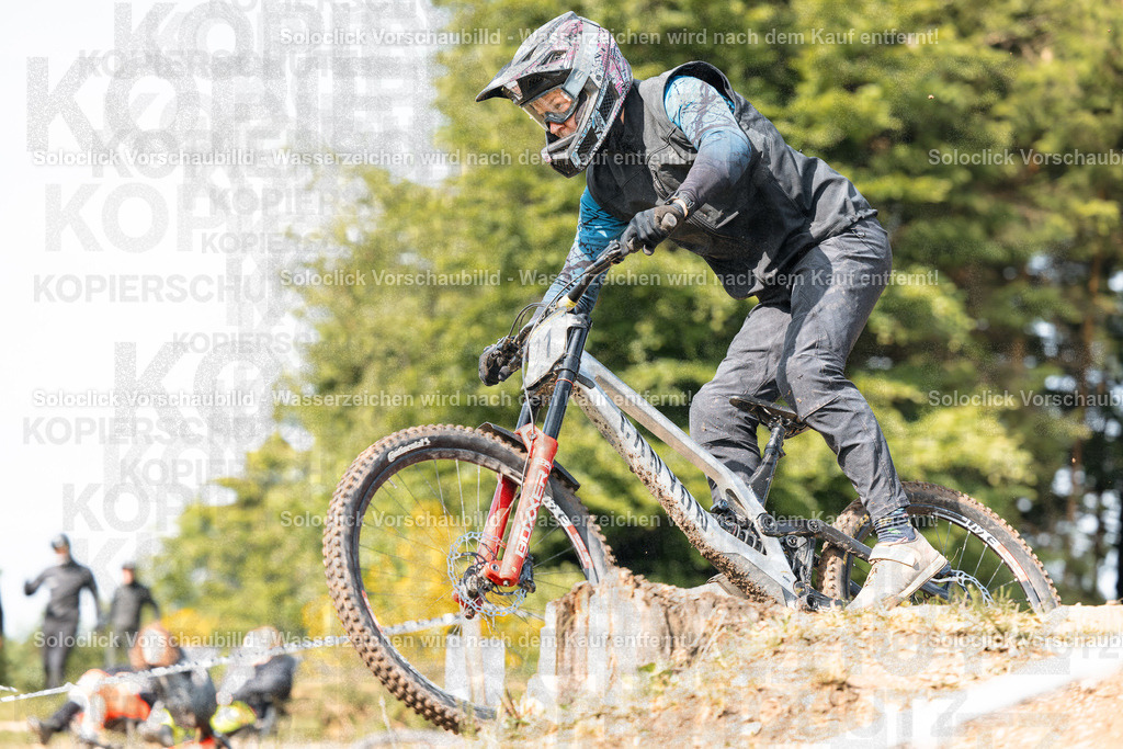 Mountainbike-Action | Ein Mountainbiker in voller Ausrüstung springt über eine kleine Erhöhung auf einem staubigen Pfad. Er trägt einen auffälligen Helm und eine passende Schutzkleidung. Der Hintergrund zeigt eine verschwommene Landschaft mit Bäumen und anderen Fahrern. - Realisiert mit Pictrs.com