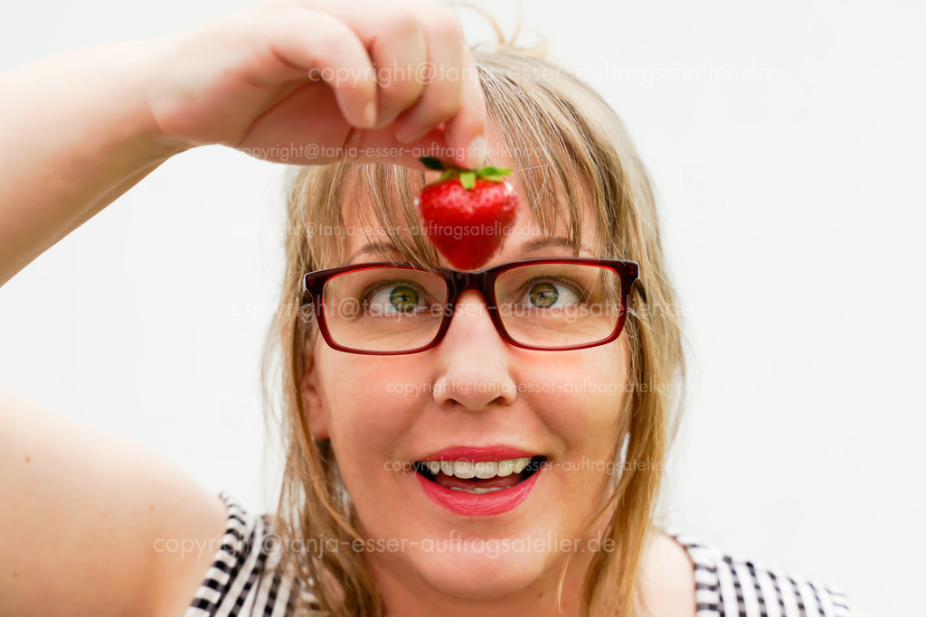 Blonde Frau hält eine Erdbeere hoch |  Eine Frau im mittleren Alter hält eine Erdbeere hoch. Sie hat blonde Haare und trägt eine Brille. Ihre Augen schauen auf die Erdbeere, deshalb schielt sie.                              